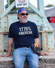 A man wearing a navy blue t-shirt with '1776% AMERICAN' printed in white, blue jeans, and a blue cap, sitting in front of a large metal bridge structure and a sign that reads "Hazard. Keep off."