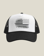 A black and white trucker hat featuring a distressed black reversed American flag graphic on the white front panel, set against a solid light beige background.