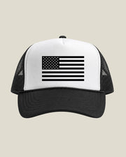 A black and white trucker hat featuring a crisp all black American flag graphic on the front white panel, set against a solid light beige background.