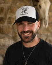 A smiling man wearing a black and white trucker hat with a crowing rooster on crossed matches graphic and the text 'YOU CAN'T SNUFF THE ROOSTER' and a black t-shirt, set against a rustic stone wall with a skull mount.