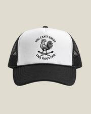 A black and white trucker hat featuring a black graphic of a crowing rooster standing on crossed matchs with the text 'YOU CAN'T SNUFF THE ROOSTER' on the front panel, set against a solid light beige background.