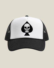 A black and white trucker hat featuring a distressed black spade with a skull graphic on the white front panel, set against a solid light beige background.
