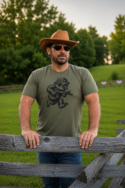 A man wearing a brown cowboy hat, aviator sunglasses, and an olive drab t-shirt with a black graphic of a sloth soldier aiming a rifle, standing behind a rustic wooden fence in a grassy field.