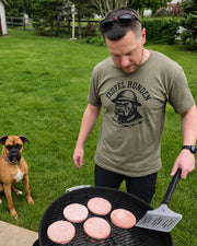 A man wearing a olive drab graphic t-shirt with a 'Teufel Hunden' devil dog logo stands next to a black grill with raw burger patties, with a brown boxer dog sitting on the grass in the background.