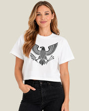 A woman wearing a white cropped boxy t-shirt with a black graphic depicting a heraldic eagle with its wings spread wide, standing on a bundle of arrows and olive branches, paired with black jeans, against a solid light beige background.