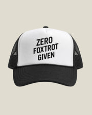 A black and white trucker hat with the text 'ZERO FOXTROT GIVEN' printed in black on the white front panel, set against a solid light beige background.